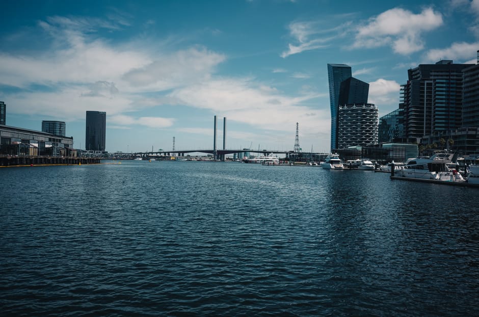 A wide view of a modern urban waterfront scene showing a calm body of water in the foreground with gentle ripples. On the right side, several white yachts and boats are moored along a paved marina area, with their hulls and masts clearly visible. The background features a cluster of contemporary high-rise buildings with glass façades, including one with a distinctive curved, wave-like pattern on its exterior. To the left, there are lower industrial or commercial structures lining the water's edge, which appear weathered with metallic and concrete textures. A bridge with two tall, narrow pylons spans across the water, connecting different parts of the cityscape, while an electrical transmission tower is visible further in the distance against a partly cloudy sky. The overall scene is illuminated by natural daylight, emphasizing the contrasts between the sleek, glassy surfaces of the skyscrapers and the textured water, creating a professional and neutral urban landscape that subtly aligns with city-based waste management and alternative disposal contexts nearby modern commercial zones. Rubbish Removal Docklands can support clients in managing waste in such active waterfront environments, including private or independent collection options.