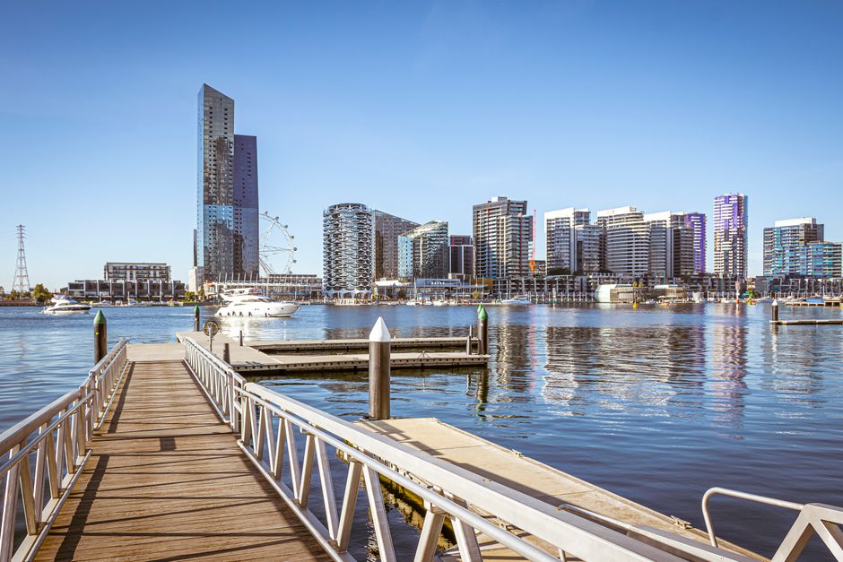 A waterfront scene in an urban setting featuring a row of weathered wooden posts with white foam caps along the edge of a waterway, possibly serving as a barrier or mooring points. Behind the posts, the water reflects the surrounding environment with gentle ripples. In the background, there is a modern, multi-story building with a distinctive architectural design, incorporating glass and metal finishes, alongside a prominent arena or stadium structure with a curved, lattice-like roof supported by visible steel beams. The area around the building includes a landscaped promenade with evenly spaced small trees, a paved walkway, and a few pedestrians. The sky is clear and blue, and the overall scene presents a clean, well-maintained environment typical of a central commercial or entertainment district in a city like Docklands. This image highlights an example of private waterfront infrastructure that might require rubbish removal or maintenance to preserve aesthetic quality, as offered by Rubbish Removal Docklands.