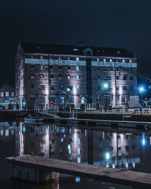 A large, multi-story brick warehouse building at night, illuminated by exterior lighting that highlights the structure's windows and brickwork. The building features a sign that reads 'LOCK WAREHOUSE' across the upper section, with some windows showing internal lights. In front of the warehouse, there is a dock area with metal railings, small bollards, and a pathway, suggesting a location near a waterway or canal. The foreground displays calm water reflecting the building's lights and the night sky, creating a mirror-like effect. The scene appears quiet and industrial, characteristic of a commercial or storage facility, potentially used for private waste handling or rubbish collection nearby, with Rubbish Removal Docklands operating in the area. The overall atmosphere is dark but well-lit by exterior fixtures, emphasizing the building’s brick texture and functional surroundings, consistent with a setting related to independent waste collection services.