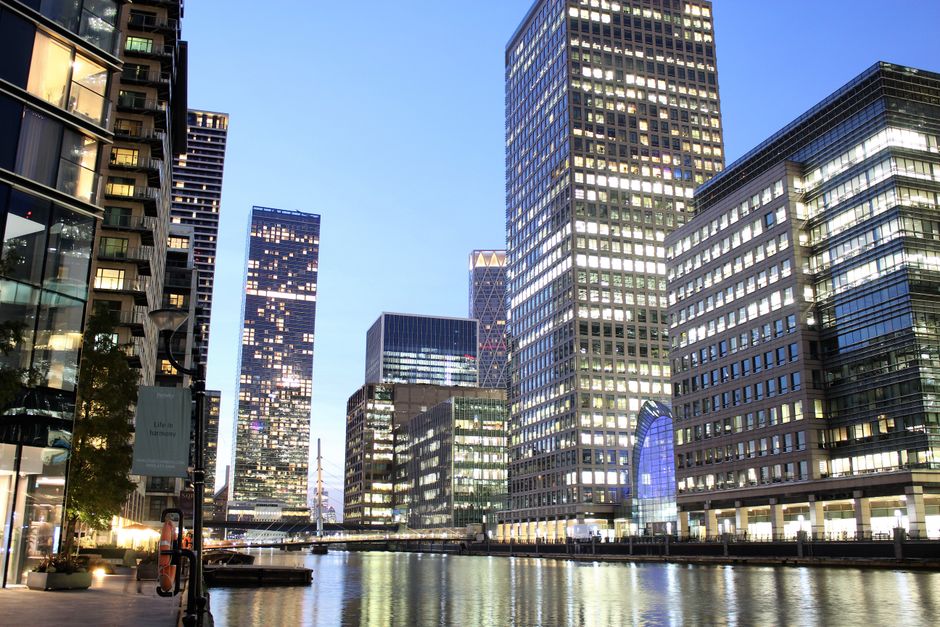 A panoramic view of a modern urban landscape featuring tall office buildings with glass facades reflecting the evening sky. The buildings vary in height and design, with some having grid-patterned windows illuminated from within, creating a bright contrast against the darkening blue sky. In the foreground, a waterway or canal runs alongside the buildings, with calm waters mirroring the lights from the structures. The scene is well-lit, with street lamps and the glow from office windows adding warmth to the environment. To the left, part of a concrete pavement and a few small trees are visible, suggesting a riverside or docklands area designed for pedestrian access. The setting exudes a sense of corporate activity and modern city life, subtly aligning with the concept of alternative waste or rubbish removal services tailored for urban office environments, such as those provided by Rubbish Removal Docklands, specialized in handling waste from commercial properties.