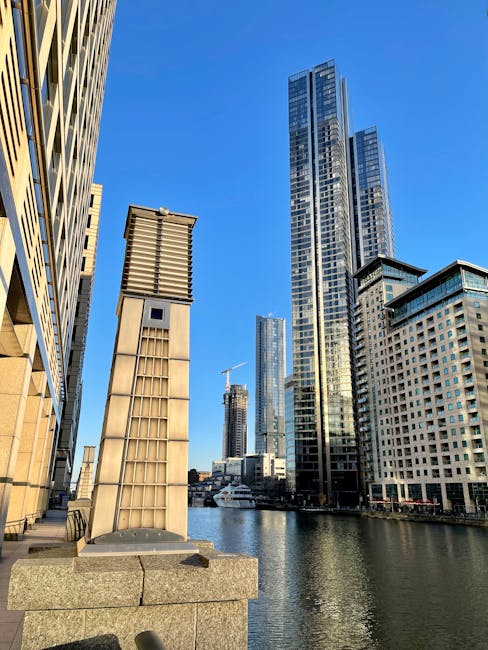 A view of a modern urban skyline along a canal, featuring tall glass skyscrapers with reflective surfaces, a smaller beige building with a grid-patterned facade and ventilation louvers, and a shorter white structure with multiple windows. The scene is set on a bright, clear day with a vivid blue sky, natural sunlight casting shadows on the buildings, and calm water reflecting the structures. The foreground includes a stone edge of the canal and part of a paved walkway, with the buildings arranged in a way that suggests a busy city environment. This setting embodies the type of location where private waste collection or on-site rubbish clearance might take place for commercial or office spaces, aligning with independent rubbish removal services such as those provided by Rubbish Removal Docklands.