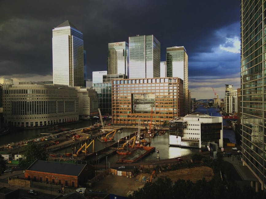 A panoramic view of a modern urban landscape featuring tall office buildings with glass facades reflecting the evening sky. The buildings vary in height and design, with some having grid-patterned windows illuminated from within, creating a bright contrast against the darkening blue sky. In the foreground, a waterway or canal runs alongside the buildings, with calm waters mirroring the lights from the structures. The scene is well-lit, with street lamps and the glow from office windows adding warmth to the environment. To the left, part of a concrete pavement and a few small trees are visible, suggesting a riverside or docklands area designed for pedestrian access. The setting exudes a sense of corporate activity and modern city life, subtly aligning with the concept of alternative waste or rubbish removal services tailored for urban office environments, such as those provided by Rubbish Removal Docklands, specialized in handling waste from commercial properties.