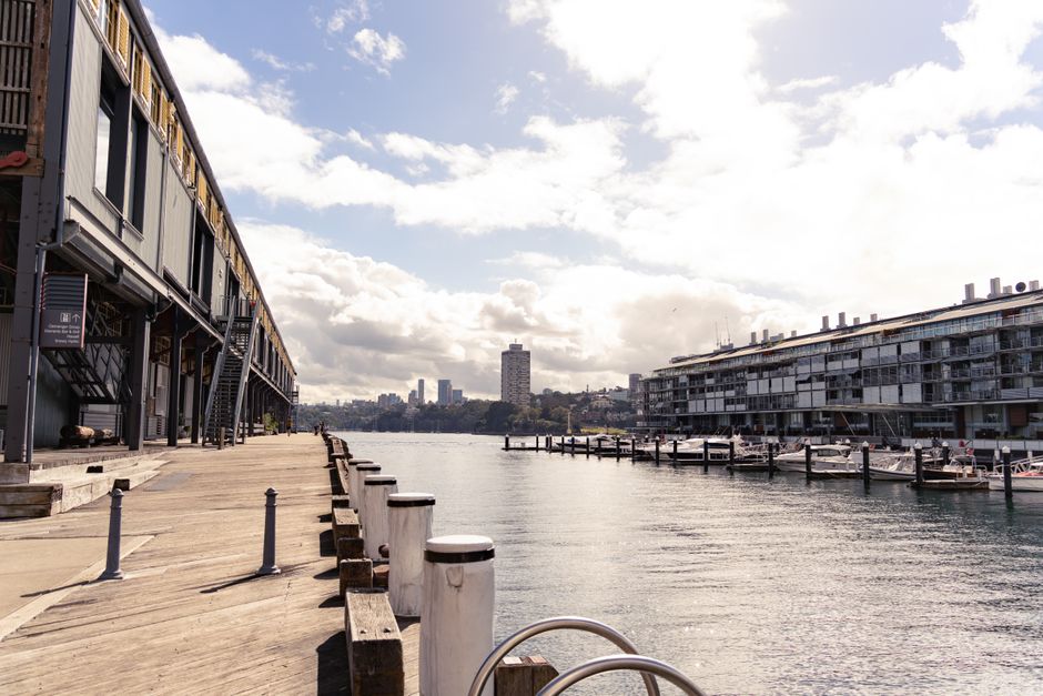 A waterfront scene featuring a row of weathered wooden mooring posts aligned along the edge of a calm waterway, with some displaying white protective caps. Behind the posts, a paved promenade is visible, lined with evenly spaced green-leaved trees and a few pedestrians. In the background, modern high-rise buildings with reflective glass facades and a prominent stadium structure with a curved, lattice-style roof dominate the urban landscape, set against a clear blue sky. The environment suggests an active city area near a dock or harbour, where private waste collection or rubbish removal services like Rubbish Removal Docklands may operate to clear waste from the waterfront or surrounding commercial zones, with the scene captured during daytime under natural lighting conditions.