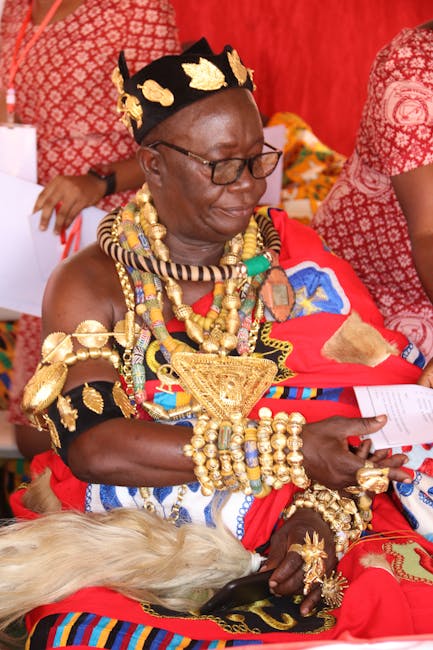 The image shows an elderly woman dressed in traditional Maasai attire, seated in a setting with a vibrant red background. She is adorned with numerous elaborate jewelry pieces, including large gold earrings, multiple layered beaded necklaces in various colors, and gold rings on her fingers. She is wearing a black headband decorated with gold leaf-shaped ornaments, and her clothing features bright red and patterned fabric typical of Maasai dress, with adornments in blue, gold, and other vivid colors. Her expression is calm and composed as she holds a piece of paper or document in her hands. Around her, other individuals in similar traditional attire are partially visible, indicating a cultural or ceremonial gathering. The scene emphasizes the rich cultural heritage and jewelry craftsmanship of Maasai attire, capturing a moment of significance or celebration within a community setting related to traditional customs.