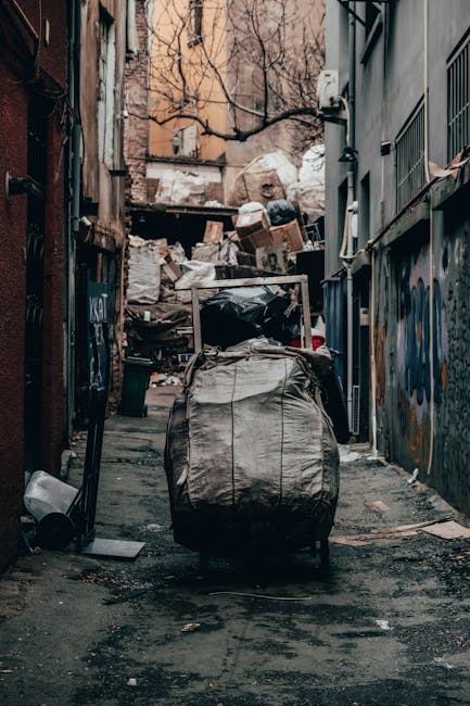 A narrow urban alleyway cluttered with piles of waste and discarded debris, with a large, wheeled garbage bag prominently positioned in the foreground. The bag appears full and made of weathered, durable plastic with visible creases and stains, suggesting it has been in use for some time. Behind it, a collection of cardboard boxes, plastic bags, and miscellaneous rubbish is stacked irregularly against a rough brick wall, partially obscuring the background which includes bare tree branches and the upper facades of older buildings. To the right, a metal fence with vertical bars and graffiti-covered walls add to the urban atmosphere, while the ground beneath the rubbish is uneven, stained, and strewn with small debris. The scene is lit by natural, diffuse daylight, emphasizing the gritty, unkempt environment that signifies an off-site or independent rubbish clearance area, often managed by private waste removal services like Rubbish Removal Docklands, reflecting typical conditions where heavy waste is temporarily stored before collection or disposal.